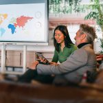 Une femme et un homme discutent en souriant sur un canapé en cuir dans un bureau. Une carte du monde est affichée à l'écran de la télé.